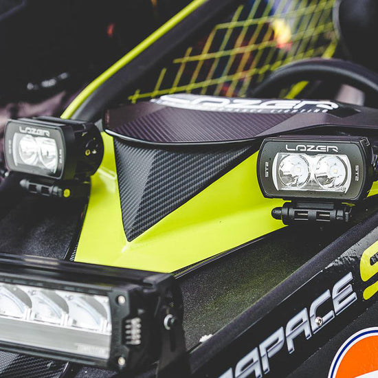 Close-up of a race car cockpit area featuring two Lozer LED light bars mounted on a bright neon yellow and black carbon-fiber vehicle body.